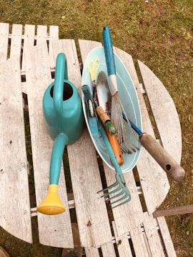 A collection of gardening tools is arranged on a wooden pallet bench outdoors. A turquoise watering can with a yellow spout is prominently placed next to a blue tray containing various gardening tools, including a pair of yellow scissors, a garden fork, and a trowel with wooden handles. The background features grass, hinting at a garden setting.