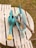 A collection of gardening tools is arranged on a wooden pallet bench outdoors. A turquoise watering can with a yellow spout is prominently placed next to a blue tray containing various gardening tools, including a pair of yellow scissors, a garden fork, and a trowel with wooden handles. The background features grass, hinting at a garden setting.