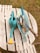 A collection of gardening tools is arranged on a wooden pallet bench outdoors. A turquoise watering can with a yellow spout is prominently placed next to a blue tray containing various gardening tools, including a pair of yellow scissors, a garden fork, and a trowel with wooden handles. The background features grass, hinting at a garden setting.