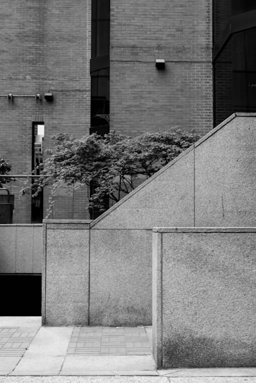 A black and white image depicting an urban environment with geometric architectural elements. The scene features a combination of brick and concrete structures, with a staircase leading upwards. Trees with foliage are visible in the background, adding a touch of nature to the otherwise stark environment.