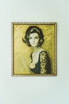 A portrait of a woman with stylized, short dark hair, wearing an elegant dress with lace sleeves and a pearl necklace. The background is textured with a soft, warm tone. The artwork is framed with an ornate golden frame.