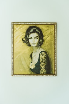 A portrait of a woman with stylized, short dark hair, wearing an elegant dress with lace sleeves and a pearl necklace. The background is textured with a soft, warm tone. The artwork is framed with an ornate golden frame.