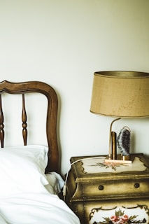 Linen upholstered nightstand beside a rustic bed, holding a vintage lamp.