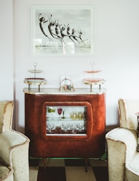 A vintage brown cabinet stands between two beige armchairs in a living space. The cabinet features a glass display with various drinking glasses. On top, there are two-tiered serving trays with decorative plates. Above the cabinet, a black-and-white photograph of people jumping is prominently displayed.