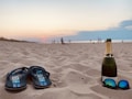A pair of flip-flops and a bottle of champagne are placed on sandy beach with sunglasses nearby. The background shows a serene sunset over the ocean with a few distant figures walking along the shoreline.