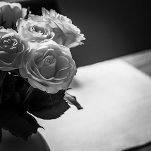 A black and white photograph of a bouquet of roses placed on a table. The flowers are positioned to the left side, with their petals and leaves clearly visible and well-defined. The background is blurred, and there is a noticeable contrast between the bright and shadowed areas in the image, giving it a dramatic feel.