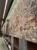 An extensive wall mural composed of various intricately designed tiles, each featuring unique patterns and designs. The mural exhibits a mixture of abstract and representational artwork with a blend of textures and colors. It stretches along a modern, well-lit corridor with smooth, reflective floors and overhead lighting.