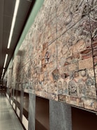 An extensive wall mural composed of various intricately designed tiles, each featuring unique patterns and designs. The mural exhibits a mixture of abstract and representational artwork with a blend of textures and colors. It stretches along a modern, well-lit corridor with smooth, reflective floors and overhead lighting.