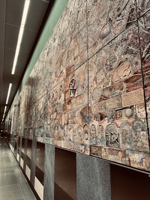 An extensive wall mural composed of various intricately designed tiles, each featuring unique patterns and designs. The mural exhibits a mixture of abstract and representational artwork with a blend of textures and colors. It stretches along a modern, well-lit corridor with smooth, reflective floors and overhead lighting.
