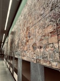 An extensive wall mural composed of various intricately designed tiles, each featuring unique patterns and designs. The mural exhibits a mixture of abstract and representational artwork with a blend of textures and colors. It stretches along a modern, well-lit corridor with smooth, reflective floors and overhead lighting.