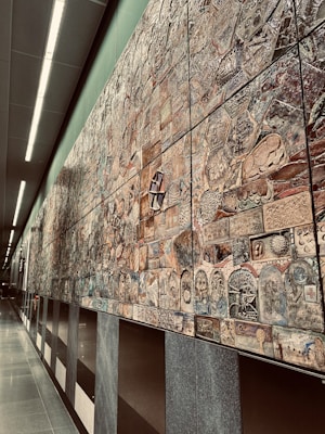 An extensive wall mural composed of various intricately designed tiles, each featuring unique patterns and designs. The mural exhibits a mixture of abstract and representational artwork with a blend of textures and colors. It stretches along a modern, well-lit corridor with smooth, reflective floors and overhead lighting.