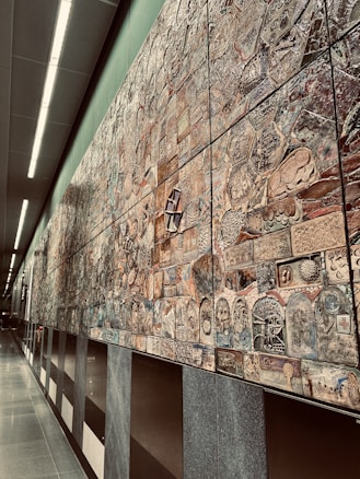 An extensive wall mural composed of various intricately designed tiles, each featuring unique patterns and designs. The mural exhibits a mixture of abstract and representational artwork with a blend of textures and colors. It stretches along a modern, well-lit corridor with smooth, reflective floors and overhead lighting.