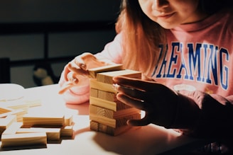 A person carefully stacking wooden blocks on a table, with a focus on their hands and the blocks. The person is wearing a pink sweater with 'DREAMING' written on it. The scene is softly lit, creating a cozy and focused atmosphere.