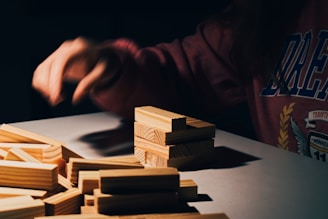 A person wearing a dark sweatshirt is seated at a table with a pile of light wooden blocks. One hand is positioned over the blocks as if in the act of placing or adjusting them. Shadows cast by the blocks create a dramatic, focused atmosphere.