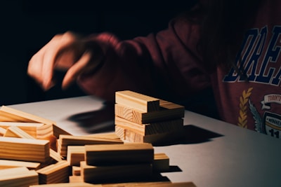 A person wearing a dark sweatshirt is seated at a table with a pile of light wooden blocks. One hand is positioned over the blocks as if in the act of placing or adjusting them. Shadows cast by the blocks create a dramatic, focused atmosphere.