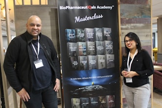 Two individuals stand on either side of a large banner for the BioPharmaceuticals Academy Masterclass. They both wear name tags and appear to be smiling. The banner lists various topics related to pharmaceuticals, including biologics and cell banks.