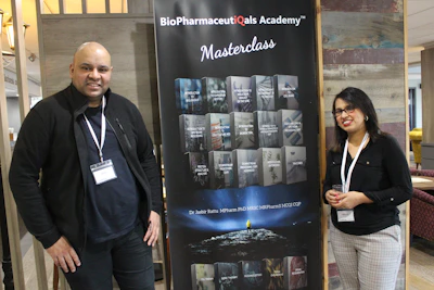 Two individuals stand on either side of a large banner for the BioPharmaceuticals Academy Masterclass. They both wear name tags and appear to be smiling. The banner lists various topics related to pharmaceuticals, including biologics and cell banks.