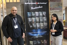 Two individuals stand on either side of a large banner for the BioPharmaceuticals Academy Masterclass. They both wear name tags and appear to be smiling. The banner lists various topics related to pharmaceuticals, including biologics and cell banks.
