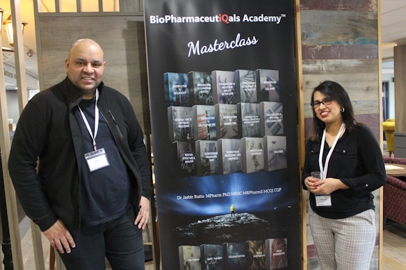Two individuals stand on either side of a large banner for the BioPharmaceuticals Academy Masterclass. They both wear name tags and appear to be smiling. The banner lists various topics related to pharmaceuticals, including biologics and cell banks.