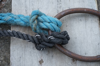 A thick blue rope and a black rope are tightly knotted around a rusty metal ring. They are lying on a wooden surface with visible grain.