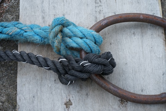 A thick blue rope and a black rope are tightly knotted around a rusty metal ring. They are lying on a wooden surface with visible grain.