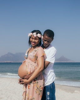 A couple stands on a beach, with the woman wearing a floral dress and a flower crown. Her pregnant belly is prominently displayed. The man stands behind her, embracing her affectionately. The ocean and mountains are visible in the background under a clear blue sky.