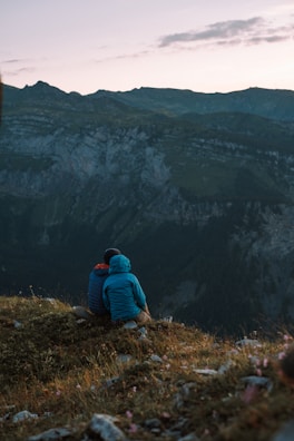 A couple resting on a mountain ridge at sunrise, wrapped in warm jackets, with misty peaks stretching behind them.