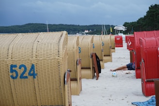 A beach scene with several wicker beach chairs, some in red and others in natural wicker color. The chairs are lined up on a sandy beach. A person is partially visible lying down next to one of the chairs, with a volleyball nearby. The background includes a forested area and a cloudy sky.