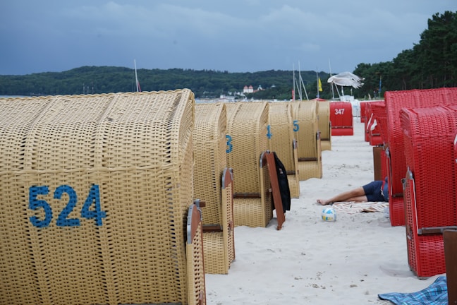 A beach scene with several wicker beach chairs, some in red and others in natural wicker color. The chairs are lined up on a sandy beach. A person is partially visible lying down next to one of the chairs, with a volleyball nearby. The background includes a forested area and a cloudy sky.