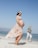 A pregnant woman in a floral dress and flower crown walks along a sandy beach. Another person in casual clothing walks behind her, smiling. The sky is clear and blue, with the ocean in the background.