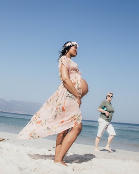 A pregnant woman in a floral dress and flower crown walks along a sandy beach. Another person in casual clothing walks behind her, smiling. The sky is clear and blue, with the ocean in the background.