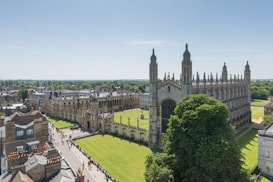 An aerial view of a large, historic Gothic-style building with an expansive courtyard covered in well-maintained green grass. The structure features intricate spires and detailed stonework, surrounded by other traditional buildings. A wide pathway with numerous people walking alongside it can be seen, leading towards the entrance. Lush trees and a clear blue sky complete the scene, suggesting a bright and sunny day.