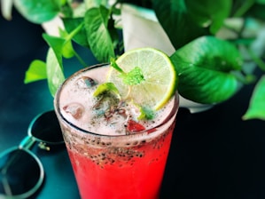 A refreshing pink drink in a clear glass filled with ice cubes and topped with chia seeds, garnished with a lime slice and mint leaves. The background is adorned with lush green plant leaves, adding a tropical feel, while a pair of sunglasses rests nearby.