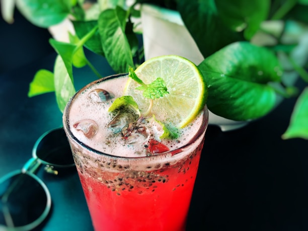 A refreshing pink drink in a clear glass filled with ice cubes and topped with chia seeds, garnished with a lime slice and mint leaves. The background is adorned with lush green plant leaves, adding a tropical feel, while a pair of sunglasses rests nearby.