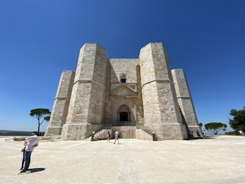 A massive stone fortress known as Castel del Monte stands prominently against a clear blue sky. The building features a unique octagonal shape with large stone towers at each corner. A lone tree is visible to the left, providing a stark contrast to the fortress's stone facade. People are standing at the base of the structure, emphasizing its grandeur and scale.