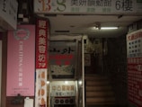 A dimly lit entrance to a building with various signs, mainly in Chinese characters. There are multiple advertising boards, some showcasing beauty services. Stairs lead up to another level, and the area looks like a narrow, commercial passage.