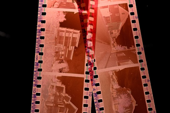 A strip of photographic film negatives with images of buildings and urban landscapes. The film features sprocket holes along its edges and has a reddish-orange tint typical of film negatives.