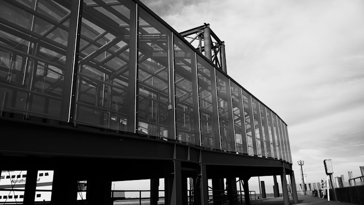A black and white image depicting a modern, glass-paneled structure with an industrial appearance. The building appears to be constructed with steel framing and large sections of glass. The architectural design features angular lines and reflects light, suggesting a sleek, contemporary style. The sky is partly cloudy, providing a contrast to the solid structure.