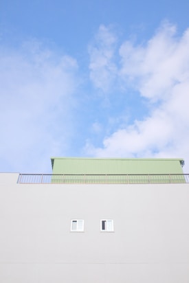 A minimalistic architectural scene features a light gray building with two small windows. Above the building is a pale green structure with a metal railing. The clear blue sky with scattered white clouds forms the backdrop.
