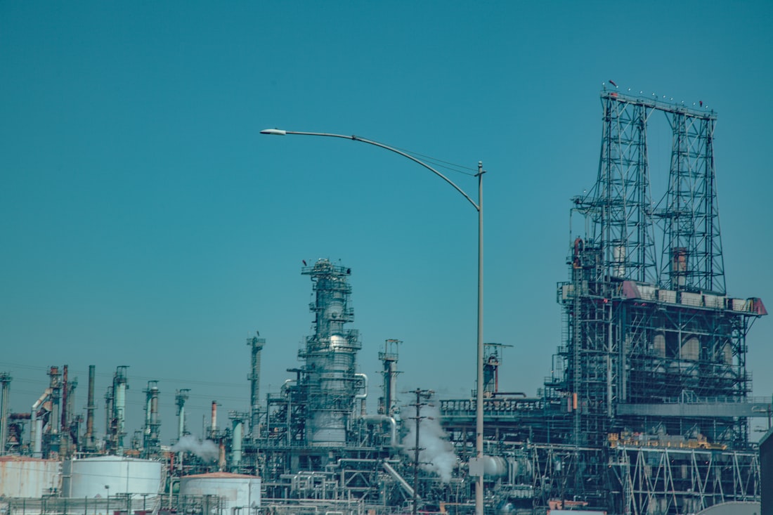 Industrial oil refinery complex under a clear blue sky — representing global energy market volatility caused by the US-Israel-Iran conflict and threats to Persian Gulf oil supply