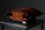 A sleek, wooden audio speaker with a unique curved shape rests on a metallic table. The speaker features rounded edges, a wood finish with a central knob, and black speaker covers. The finish has a rich, dark wood grain accented by metallic elements.