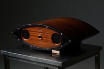 A sleek, wooden audio speaker with a unique curved shape rests on a metallic table. The speaker features rounded edges, a wood finish with a central knob, and black speaker covers. The finish has a rich, dark wood grain accented by metallic elements.