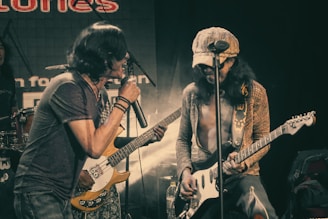 Two musicians perform passionately on stage. One is singing into a microphone while holding a bass guitar, and the other plays an electric guitar. The scene is vibrant with stage lights and a visible backdrop displaying partial text.