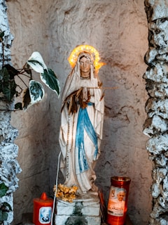 A religious statue of the Virgin Mary adorned with a glowing halo stands inside a stone alcove. The figure is dressed in white and blue garments and is surrounded by prayer candles, one of which features an image of a religious figure. Small decorative plants and flowers are placed around the statue, enhancing the sacred ambiance.