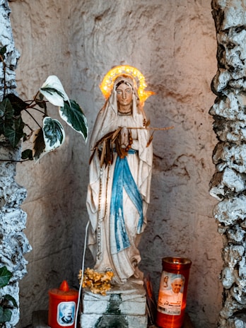 A religious statue of the Virgin Mary adorned with a glowing halo stands inside a stone alcove. The figure is dressed in white and blue garments and is surrounded by prayer candles, one of which features an image of a religious figure. Small decorative plants and flowers are placed around the statue, enhancing the sacred ambiance.