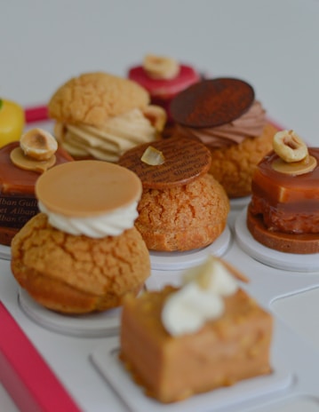 An assortment of colorful pastries is arranged, each topped with various ingredients like cream, chocolate disks, and hazelnuts. The pastries feature a range of textures, including smooth, creamy, and crumbly surfaces. They rest on a white tray with a pink border.