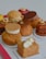 An assortment of colorful pastries is arranged, each topped with various ingredients like cream, chocolate disks, and hazelnuts. The pastries feature a range of textures, including smooth, creamy, and crumbly surfaces. They rest on a white tray with a pink border.