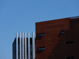 The image features a modern architectural building with a section of red-brown cladding on the right and a series of metallic cylindrical structures on the left. The sky is clear and blue, adding a stark contrast to the industrial elements of the building.