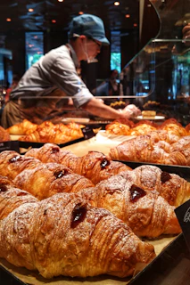 A person wearing a cap and mask is working behind a glass display filled with freshly baked croissants. The pastries have a golden-brown crust with visible layers and some have a dollop of jelly on top. The setting appears to be a bakery with warm lighting and a variety of baked goods on display.