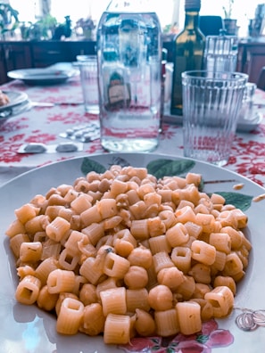 A delicious plate features pasta e ceci, a simple Italian dish made with pasta and chickpeas, placed on a floral-decorated plate. The background reveals a dining table set with a bottle of water, an olive oil bottle, and several glasses, all atop a tablecloth adorned with red patterns.